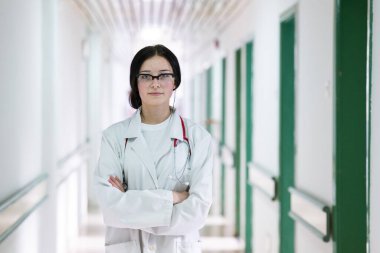 Kendine güvenen, steteskoplu genç bir bayan doktor. Parlak bir hastane koridorunda dururken gülümsüyor.. 