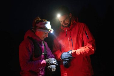 Gece yürüyüşçüleri, düşük ışık koşulları altında navigasyon ve takım çalışmasını vurgulayan far ışığı altında bir cihazı incelerler..