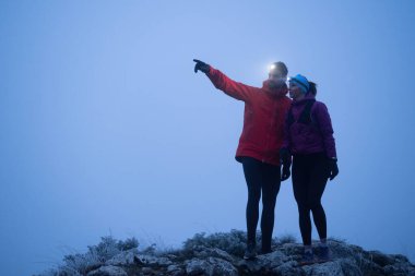 İki yürüyüşçü, sabahın erken saatlerinde bir dağın zirvesinde, etrafı sisle çevrili farlarla GPS cihazlarına danışırlar..