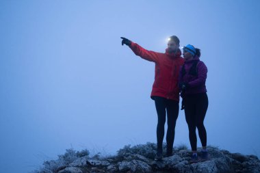 İki yürüyüşçü, sabahın erken saatlerinde bir dağın zirvesinde, etrafı sisle çevrili farlarla GPS cihazlarına danışırlar..