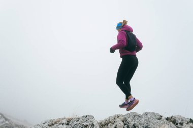 Soğuk ve sisli havada kayalık bir dağ yolunda koşan bir kadın sporcu, sıcak bir ceket, eldiven ve saç bandı giyiyor, dayanıklılık ve kararlılık sergiliyor.. 