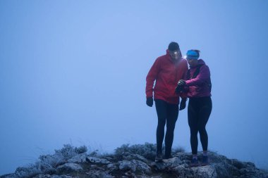 İki yürüyüşçü, sabahın erken saatlerinde bir dağın zirvesinde, etrafı sisle çevrili farlarla GPS cihazlarına danışırlar..