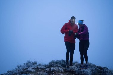 İki yürüyüşçü, sabahın erken saatlerinde bir dağın zirvesinde, etrafı sisle çevrili farlarla GPS cihazlarına danışırlar..