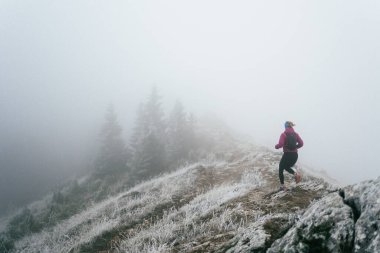 Soğuk ve sisli havada kayalık bir dağ yolunda koşan bir kadın sporcu, sıcak bir ceket, eldiven ve saç bandı giyiyor, dayanıklılık ve kararlılık sergiliyor.. 