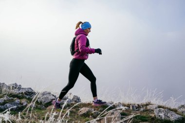 Soğuk ve sisli havada kayalık bir dağ yolunda koşan bir kadın sporcu, sıcak bir ceket, eldiven ve saç bandı giyiyor, dayanıklılık ve kararlılık sergiliyor.. 
