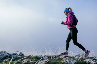Soğuk ve sisli havada kayalık bir dağ yolunda koşan bir kadın sporcu, sıcak bir ceket, eldiven ve saç bandı giyiyor, dayanıklılık ve kararlılık sergiliyor.. 