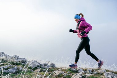 Soğuk ve sisli havada kayalık bir dağ yolunda koşan bir kadın sporcu, sıcak bir ceket, eldiven ve saç bandı giyiyor, dayanıklılık ve kararlılık sergiliyor.. 
