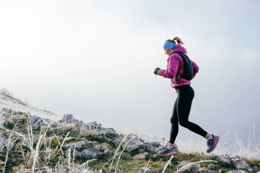 Soğuk ve sisli havada kayalık bir dağ yolunda koşan bir kadın sporcu, sıcak bir ceket, eldiven ve saç bandı giyiyor, dayanıklılık ve kararlılık sergiliyor.. 