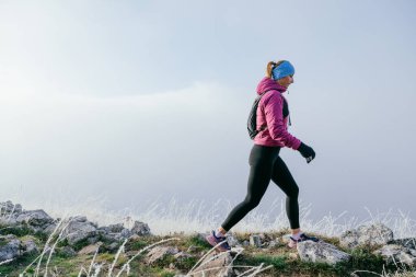 Soğuk ve sisli havada kayalık bir dağ yolunda koşan bir kadın sporcu, sıcak bir ceket, eldiven ve saç bandı giyiyor, dayanıklılık ve kararlılık sergiliyor.. 