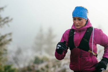 Soğuk ve sisli havada kayalık bir dağ yolunda koşan bir kadın sporcu, sıcak bir ceket, eldiven ve saç bandı giyiyor, dayanıklılık ve kararlılık sergiliyor.. 