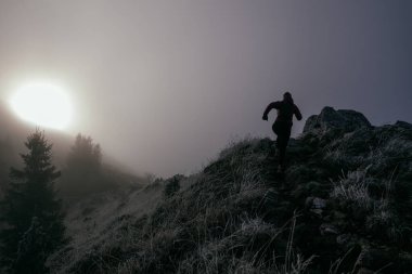Soğuk ve sisli havada kayalık bir dağ yolunda koşan bir kadın sporcu, sıcak bir ceket, eldiven ve saç bandı giyiyor, dayanıklılık ve kararlılık sergiliyor.. 