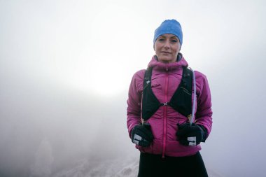 Pembe ceketli, kendine güveni tam bir kadın yürüyüşçü sisli bir dağın zirvesinde duruyor..