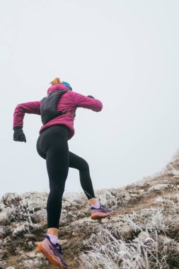 Soğuk ve sisli havada kayalık bir dağ yolunda koşan bir kadın sporcu, sıcak bir ceket, eldiven ve saç bandı giyiyor, dayanıklılık ve kararlılık sergiliyor.. 