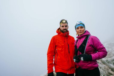 Açık hava kıyafetleri içinde sisli bir dağın tepesinde poz veren iki yürüyüşçünün portresi.