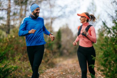 Gülümseyen bir erkek ve kadın, serin bir günde, doğa, fitness ve birbirlerine eşlik ederek, spor giysileri içinde, manzaralı bir orman patikasında birlikte koşarlar..