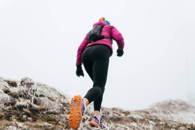 Soğuk ve sisli havada kayalık bir dağ yolunda koşan bir kadın sporcu, sıcak bir ceket, eldiven ve saç bandı giyiyor, dayanıklılık ve kararlılık sergiliyor.. 