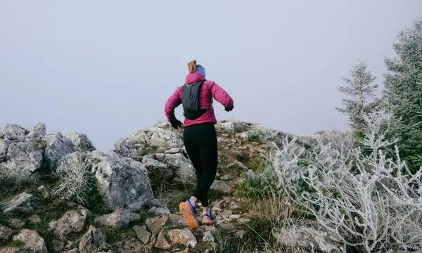 Soğuk ve sisli havada kayalık bir dağ yolunda koşan bir kadın sporcu, sıcak bir ceket, eldiven ve saç bandı giyiyor, dayanıklılık ve kararlılık sergiliyor.. 