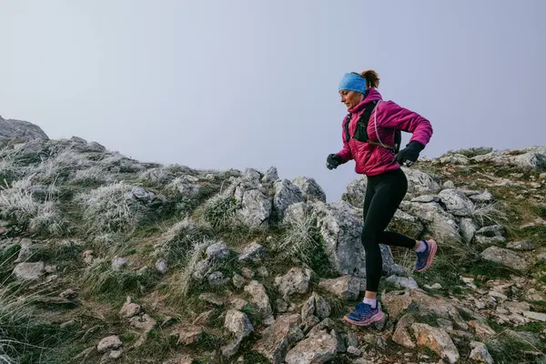 Soğuk ve sisli havada kayalık bir dağ yolunda koşan bir kadın sporcu, sıcak bir ceket, eldiven ve saç bandı giyiyor, dayanıklılık ve kararlılık sergiliyor..
