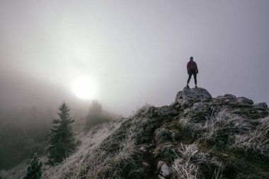 Soğuk ve sisli havada kayalık bir dağ yolunda koşan bir kadın sporcu, sıcak bir ceket, eldiven ve saç bandı giyiyor, dayanıklılık ve kararlılık sergiliyor.. 