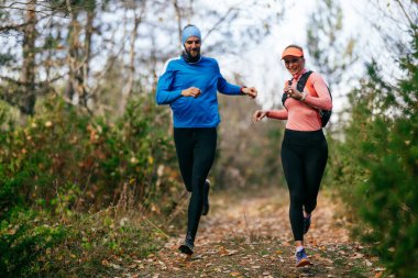Gülümseyen bir erkek ve kadın, serin bir günde, doğa, fitness ve birbirlerine eşlik ederek, spor giysileri içinde, manzaralı bir orman patikasında birlikte koşarlar..
