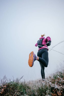 Pembe ceketli, siyah taytlı, yokuş yukarı koşan kadın koşucunun arka görüntüsü.