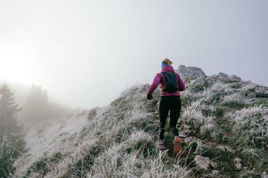 Soğuk ve sisli havada kayalık bir dağ yolunda koşan bir kadın sporcu, sıcak bir ceket, eldiven ve saç bandı giyiyor, dayanıklılık ve kararlılık sergiliyor.. 