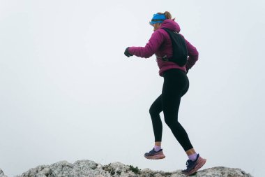 Soğuk ve sisli havada kayalık bir dağ yolunda koşan bir kadın sporcu, sıcak bir ceket, eldiven ve saç bandı giyiyor, dayanıklılık ve kararlılık sergiliyor.. 