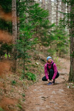 Koşan ayakkabısını bağlamak için çömelmiş kadın yürüyüşçü çam ağaçlarıyla çevrili bir orman yoluna, spor kış kıyafetleri içinde..