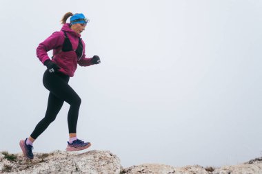 Soğuk ve sisli havada kayalık bir dağ yolunda koşan bir kadın sporcu, sıcak bir ceket, eldiven ve saç bandı giyiyor, dayanıklılık ve kararlılık sergiliyor.. 