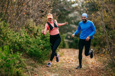 Gülümseyen bir erkek ve kadın, serin bir günde, doğa, fitness ve birbirlerine eşlik ederek, spor giysileri içinde, manzaralı bir orman patikasında birlikte koşarlar..