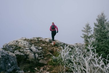 Soğuk ve sisli havada kayalık bir dağ yolunda koşan bir kadın sporcu, sıcak bir ceket, eldiven ve saç bandı giyiyor, dayanıklılık ve kararlılık sergiliyor.. 