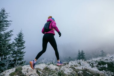 Soğuk ve sisli havada kayalık bir dağ yolunda koşan bir kadın sporcu, sıcak bir ceket, eldiven ve saç bandı giyiyor, dayanıklılık ve kararlılık sergiliyor.. 