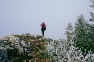 Soğuk ve sisli havada kayalık bir dağ yolunda koşan bir kadın sporcu, sıcak bir ceket, eldiven ve saç bandı giyiyor, dayanıklılık ve kararlılık sergiliyor.. 