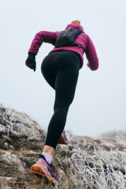Soğuk ve sisli havada kayalık bir dağ yolunda koşan bir kadın sporcu, sıcak bir ceket, eldiven ve saç bandı giyiyor, dayanıklılık ve kararlılık sergiliyor.. 