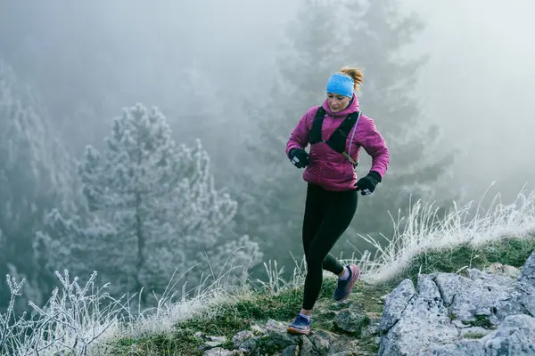 Soğuk ve sisli havada kayalık bir dağ yolunda koşan bir kadın sporcu, sıcak bir ceket, eldiven ve saç bandı giyiyor, dayanıklılık ve kararlılık sergiliyor.. 
