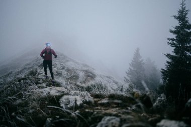 Soğuk ve sisli havada kayalık bir dağ yolunda koşan bir kadın sporcu, sıcak bir ceket, eldiven ve saç bandı giyiyor, dayanıklılık ve kararlılık sergiliyor.. 
