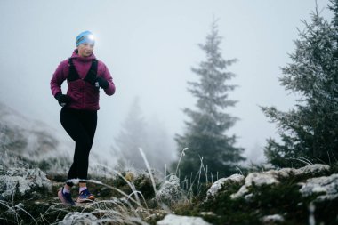 Soğuk ve sisli havada kayalık bir dağ yolunda koşan bir kadın sporcu, sıcak bir ceket, eldiven ve saç bandı giyiyor, dayanıklılık ve kararlılık sergiliyor.. 