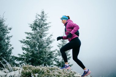 Soğuk ve sisli havada kayalık bir dağ yolunda koşan bir kadın sporcu, sıcak bir ceket, eldiven ve saç bandı giyiyor, dayanıklılık ve kararlılık sergiliyor.. 
