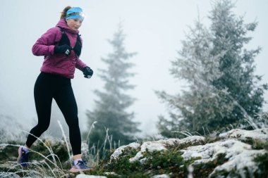 Soğuk ve sisli havada kayalık bir dağ yolunda koşan bir kadın sporcu, sıcak bir ceket, eldiven ve saç bandı giyiyor, dayanıklılık ve kararlılık sergiliyor.. 
