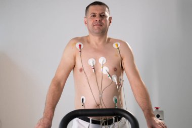 Orta yaşlı bir adam, klinik bir ortamda kalp izleme için EKG elektrotlarına bağlı bir koşu bandında tıbbi stres testinden geçiyor.. 