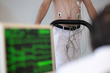 Orta yaşlı bir adam, klinik bir ortamda kalp izleme için EKG elektrotlarına bağlı bir koşu bandında tıbbi stres testinden geçiyor.. 