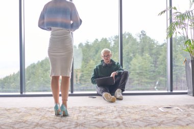 Sarışın bir adam modern bir ofisin zemininde rastgele oturuyor, dizüstü bilgisayarını kullanırken pencereye yaslanıyor, yanında çekici bayan arkadaşı duruyor..