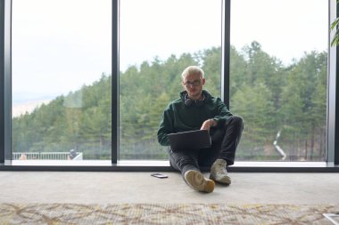 Modern bir ofisin zemininde oturan sarışın bir adam, kucağında dizüstü bilgisayarla pencereye yaslanmış, rahat ve odaklanmış bir tavır sergilerken tamamen işine odaklanmış.