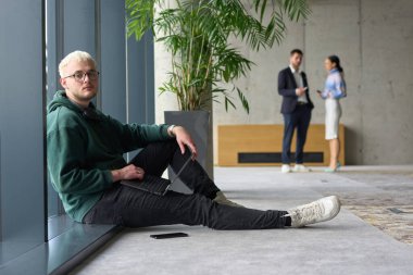 Modern bir ofiste geniş bir pencerenin yanında oturan sıradan bir profesyonel bilgisayarla çalışırken gülümseyen meslektaşları bulanık arka planda etkileşime giren biri.