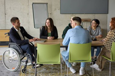 Muhtelif bir grup iş adamı, yöneticileriyle bir ofiste oturuyorlar, şu anda tekerlekli sandalyede, işbirliği, liderlik ve kapsayıcılığı destekliyorlar..