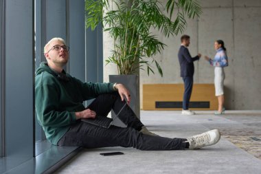 Modern bir ofiste geniş bir pencerenin yanında oturan sıradan bir profesyonel bilgisayarla çalışırken gülümseyen meslektaşları bulanık arka planda etkileşime giren biri.