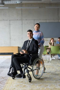 Profesyonel bir ofis ortamında, bir iş kadını tekerlekli sandalyede oturan ve tablet kullanan yönetmenine yardım ederken, meslektaşları arka planda işbirliği yapıyor.