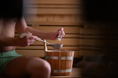 Geleneksel ahşap bir saunanın içinde elinde kova ve kepçeyle gerekli yağları tutan bir adamın eli yakındır..