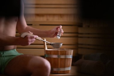 Geleneksel ahşap bir saunanın içinde elinde kova ve kepçeyle gerekli yağları tutan bir adamın eli yakındır..