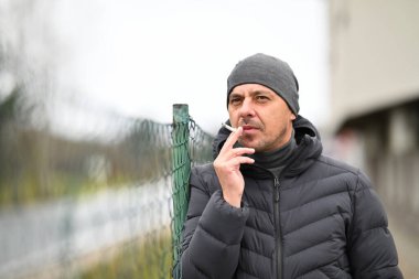 Kışlık ceketli ve bereli bir adam dışarıdaki tel örgülerin yanında sigara içiyor..